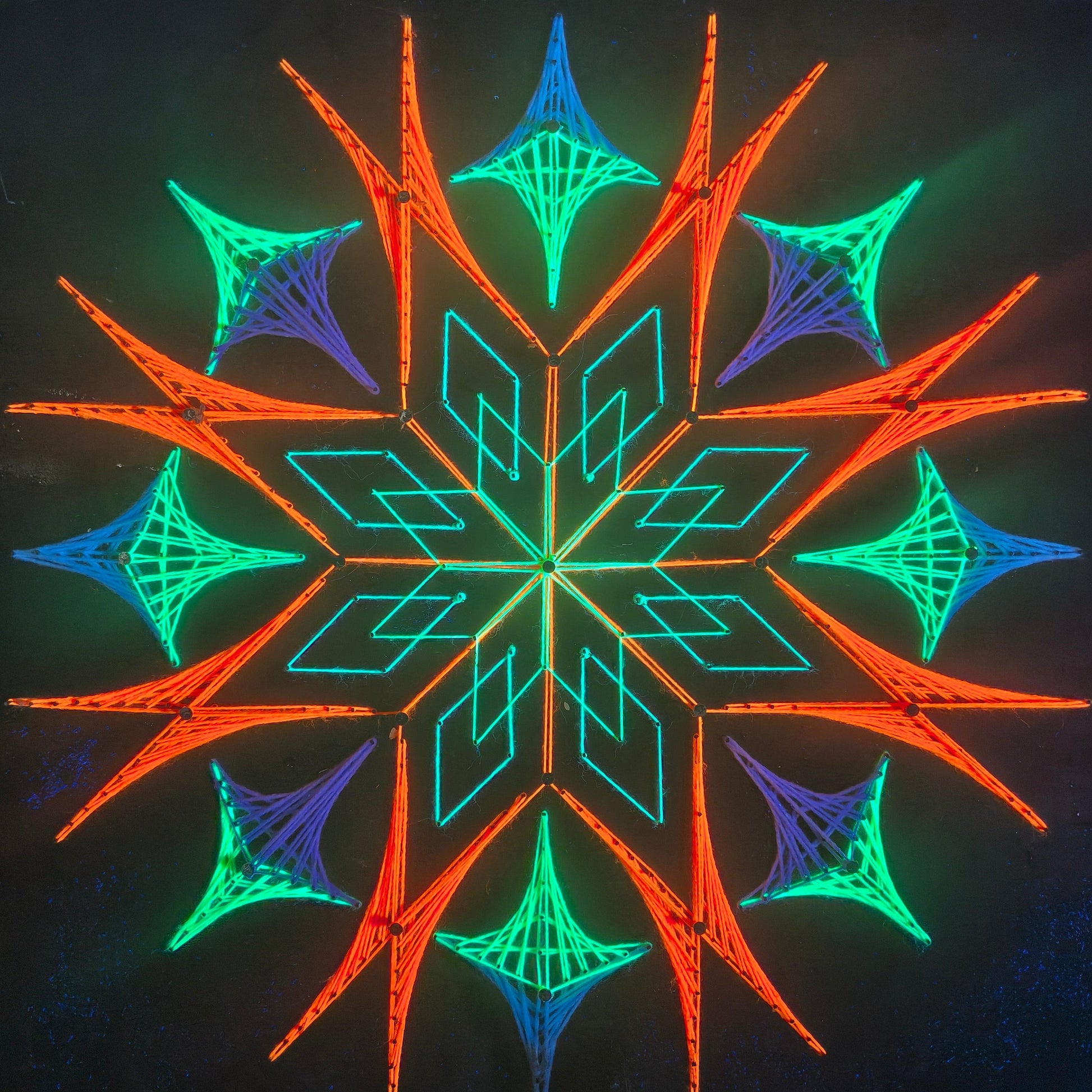 handmade UV reactive square string art with symmetrical flame-inspired geometric pattern, neon orange, green and blue threads, glowing under ultraviolet light, by Psybell
