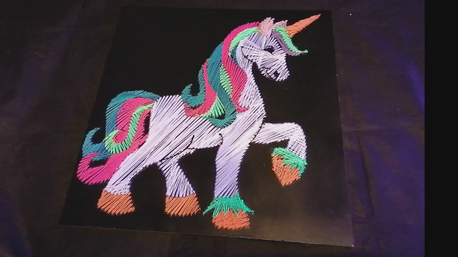 handmade UV reactive unicorn string art made from neon cotton threads, glowing under ultraviolet light, geometric silhouette with rainbow mane and tail on black MDF board by Psybell