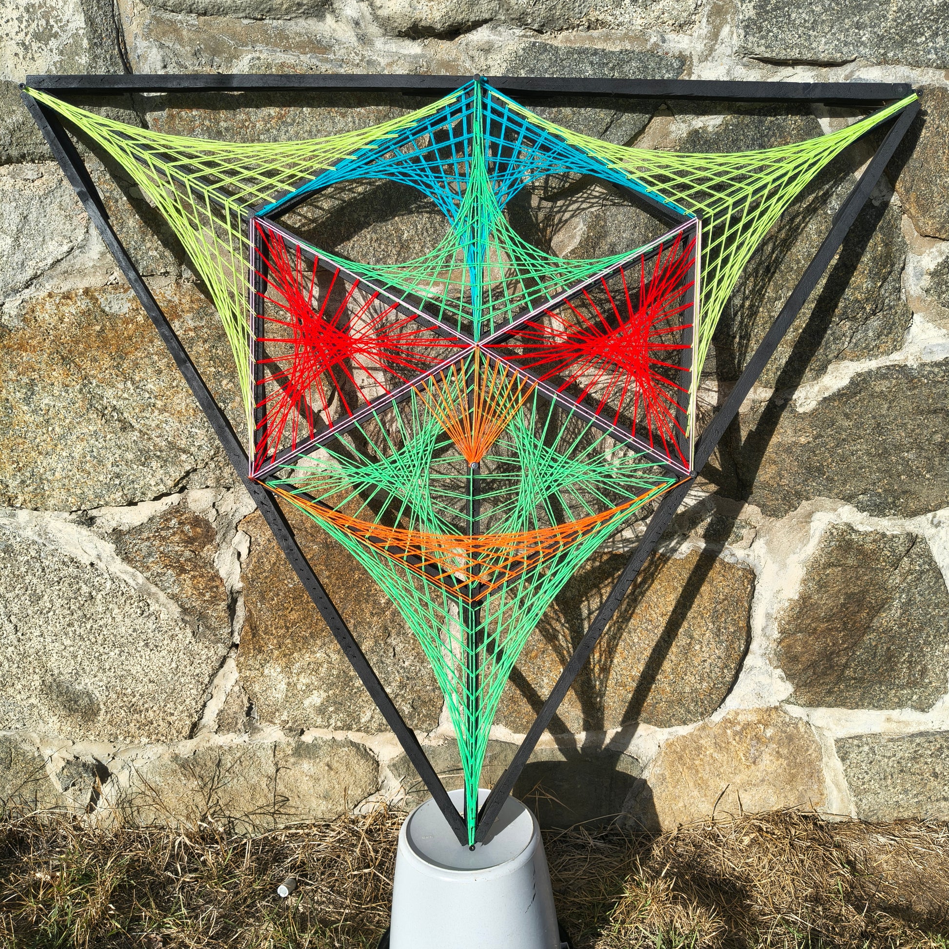 Geometric string art wall sculpture Alien Hunter by Psybell displayed outdoors against a stone wall