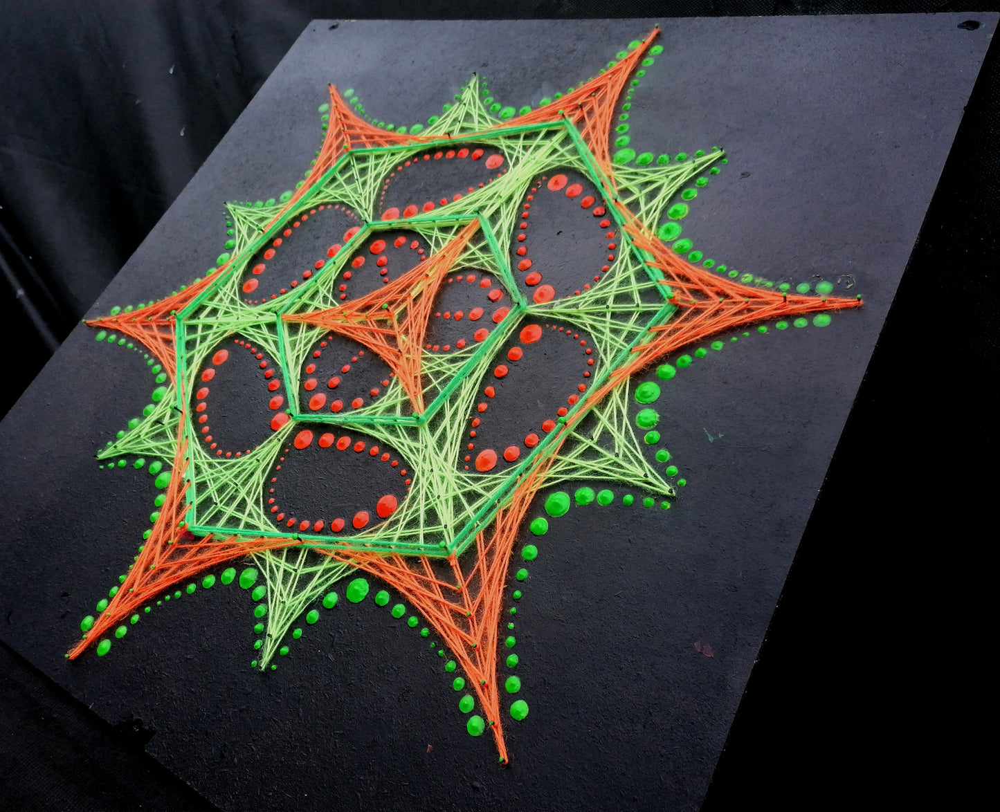 Angled view of UV reactive dotted mandala string art “Transformer” by Psybell with neon threads on black board